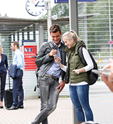 Zu sehen sind zwei Menschen welche an einer Haltestelle stehen und auf ein Smartphone schauen.